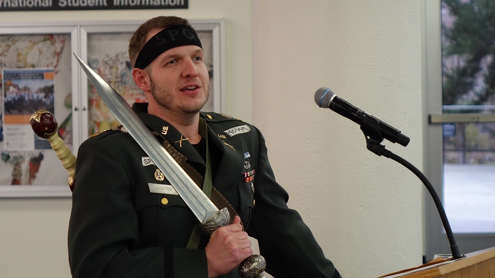 Man speaking at a podium holding a sword on his shoulder