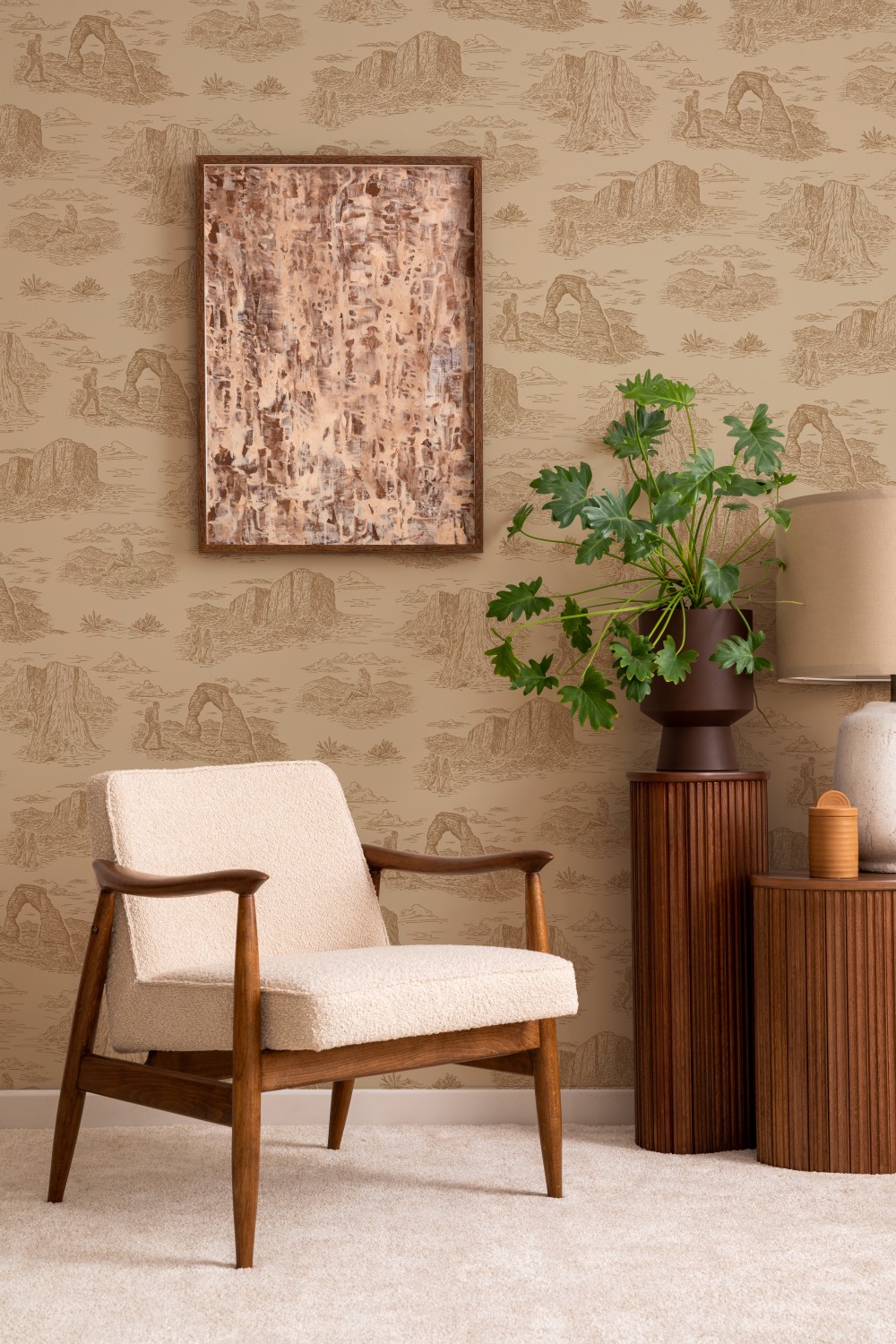 A vignette with a chair and accent tables with a plant on top and a beige and burnt orange
wallpaper behind it featuring images of Delicate Arch, red cliffs, and vegetation local to
Southern Utah.